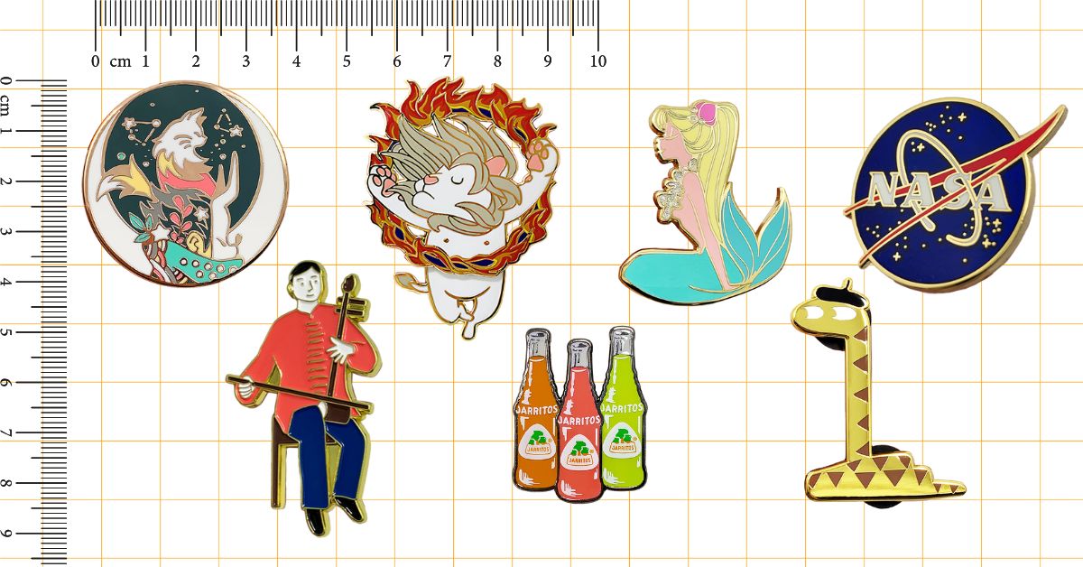 A collection of custom enamel pins with various designs shown next to a ruler for size reference.